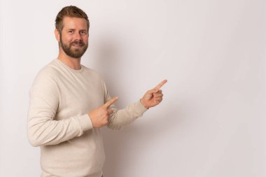 Süveter giyen genç bir adam, bir ürünü ya da bir fikri sunmak için parmağıyla kenarı gösterirken izole edilmiş beyaz arka planda gülümseyerek ileriye bakıyor.