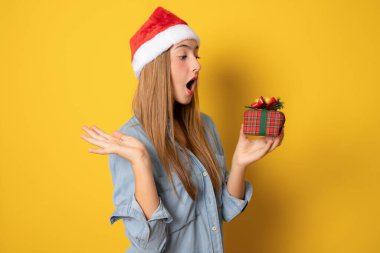 Sarı arka planda izole edilmiş Noel Baba şapkalı genç bir kadın hediye kutusunu tutuyor..