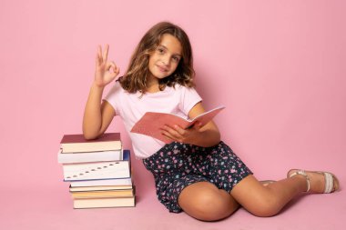 Güzel küçük kız yere oturup kitap okuyor. Pembe arka planda izole edilmiş