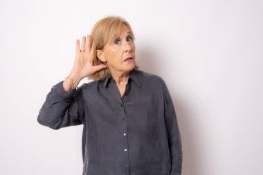 Mavi gömlekli neşeli yaşlı kadının portresi beyaz arka planda izole edilmiş bir şekilde dinleme hareketi yapıyor.