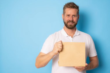 Genç gülümseyen bir adam açık kahverengi boş el işi kağıt paket paketi gösteriyor. Mavi arka planda izole edilmiş gibi duruyor..