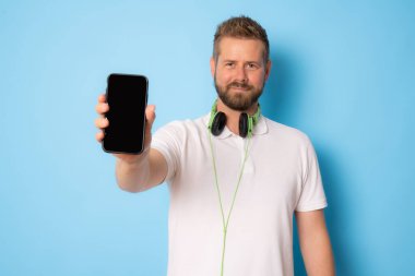 Mavi arka planda izole edilmiş günlük giysiler giyen neşeli bir gencin portresi, boş ekranlı cep telefonunu gösteriyor.
