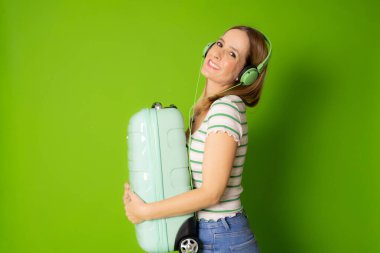 Kulaklıklı genç bir kadın yeşil arka planda izole edilmiş bavulu tutuyor..