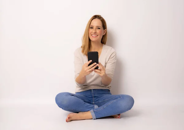 Uśmiechnięta Kobieta Pomocą Smartfona Patrząc Aparatu Siedząc Podłodze Skrzyżowanymi Nogami — Zdjęcie stockowe