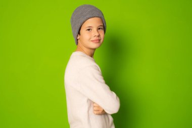 Kışın giyinmiş, kolları kapalı yeşil arka planda kameraya bakan sevimli genç bir çocuğun fotoğrafı.