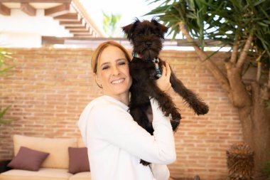 Gülümseyen genç bir kadın dışarıda köpeğine sarılıyor..