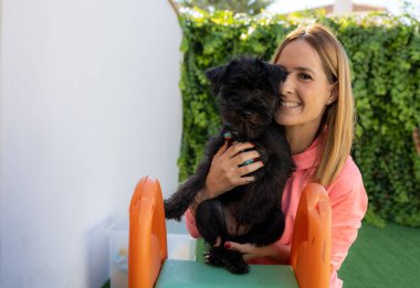 Açık havada köpeğini kucaklayan gülümseyen genç bir kadın..