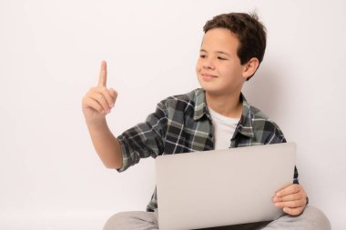 Ekose gömlek giyen, elinde dizüstü bilgisayarla oturan ve beyaz arka planda izole edilmiş bir şekilde parmağını kaldıran sevimli bir çocuk..