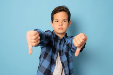 Mavi arka planda ekose gömlek giyen sevimli beyaz çocuk kızgın, sinirli, hayal kırıklığına uğramış ya da hoşnutsuz, ciddi bir bakışla başparmağını kaldırıyor.