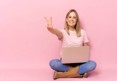 Dizüstü bilgisayar kullanan çizgili tişörtlü genç bir kadın pembe arka planda barış sembolü olarak oturuyor..