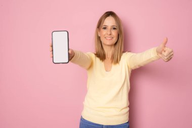 Genç, gülümseyen bir kadın pembe arka planda parmaklarını kaldırarak cep telefonu ekranı gösteriyor..