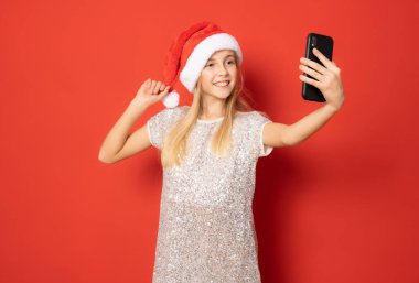 Gülümseyen kız Noel Baba şapkası takıyor. Akıllı telefon kullanıyor. Kırmızı arka planda tek başına duruyor..