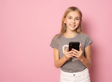 Gülümseyen, yaz tişörtlü, pembe arka planda cep telefonu kullanan küçük bir kız..