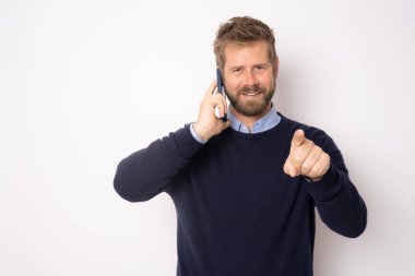 Genç, gülümseyen, cep telefonuyla konuşan, kamerayı işaret eden, beyaz arka planda tek başına duran yakışıklı adam..