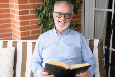 Kıdemli adam evde terasta otururken rahatlamak için güzel bir kitabın tadını çıkarıyor..