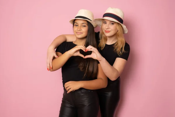 两个戴黑帽和草帽的女孩在一起摆出一副心形 手指与粉色背景隔离在一起 — 图库照片