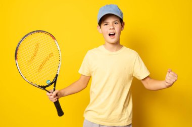 Sırıtkan çocuk, sarı arka planda tenisçi raketi tutuyor.