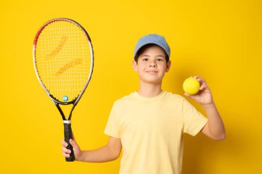 Sırıtkan çocuk, sarı arka planda tenisçi raketi tutuyor.