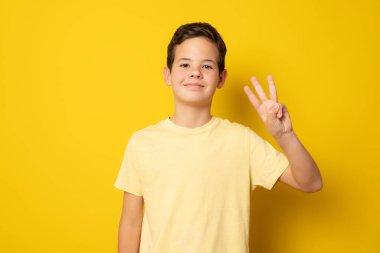 Sıradan tişörtlü gülümseyen çocuk sarı arka planda parmakları izole edilmiş üç kişi sayıyor..