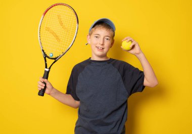Sırıtkan çocuk, sarı arka planda tenisçi raketi tutuyor.