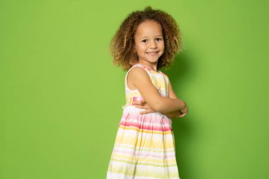 Çekici, sevimli, küçük bir kızın fotoğrafı. Elleri katlanmış, yeşil arka planda duruyor..
