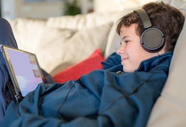 Evdeki kanepede kulaklık takıp tablet kullanan sevimli çocuk.