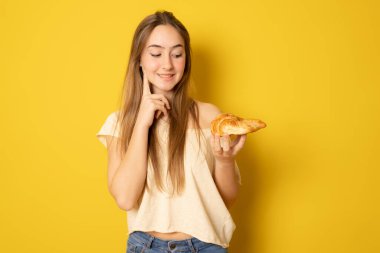 Young woman eating croissant, coffee break. Isolated over yellow background. Diet concept.