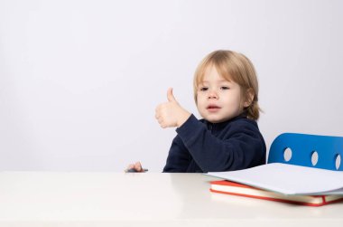 Masada oturan küçük çocuk beyaz arka planda izole bir şekilde baş parmağını kaldırıyor..