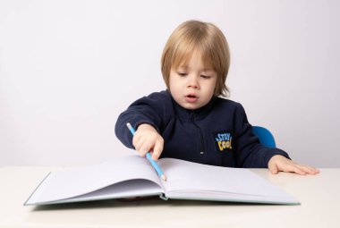 Küçük çocuk masada oturuyor ve beyaz bir arka plan çiziyor.