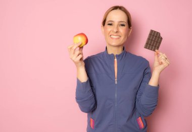 Kafası karışmış genç bir kadının portresi. Pembe arka plandan izole edilmiş çikolata ve yeşil elma arasında seçim yapıyor.