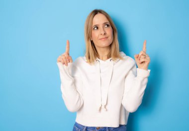 Gülümseyen genç kadın, günlük giysiler içinde parmaklarını havaya kaldırarak mavi arka planda izole bir şekilde duruyor..