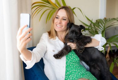 Evde sevimli köpeğiyle cep telefonu kullanan gülümseyen genç bir kadın..