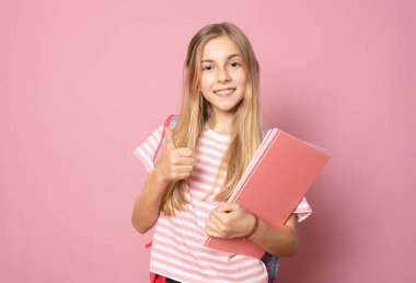 Sırt çantası takan genç ve güzel bir kız pembe arka planda tek başına başparmağını kaldırmış duruyor. Eğitim kavramı.
