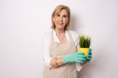 Bahçe işleri, tarım ve yaşlılar konsepti. Önlüklü gülümseyen yaşlı bir kadının portresi. Beyaz arka planda izole edilmiş küçük bir bitki tutuyor..