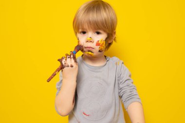 Güzel küçük çocuk masada oturuyor ve sarı arka planda boya ve fırçalarla renkli resimler çiziyor. Çocuk konsepti.