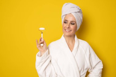 Bornozlu genç mutlu kadın, sarı arka planda tek başına dikilirken cilt bakımı için yeşim taşı kullanıyor. Güzellik konsepti.