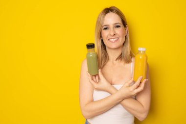 Günlük giysiler içinde gülümseyen genç bir kadın sarı arka planda izole edilmiş detoks diyeti olarak sıkılmış yeşil turuncu sebze suyu tutuyor. İnsanların yaşam tarzı gıda konsepti