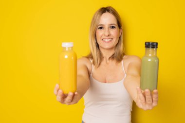 Günlük giysiler içinde gülümseyen genç bir kadın sarı arka planda izole edilmiş detoks diyeti olarak sıkılmış yeşil turuncu sebze suyu tutuyor. İnsanların yaşam tarzı gıda konsepti
