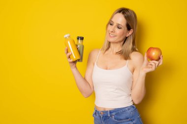 Günlük giysiler içinde gülümseyen genç bir kadın sarı arka planda izole edilmiş detoks diyeti olarak sıkılmış yeşil turuncu sebze suyu tutuyor. İnsanların yaşam tarzı gıda konsepti