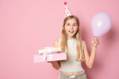 Doğum günü şapkalı neşeli küçük bir kızın portresi pembe arka planda izole edilmiş hediye kutularını gösteriyor.