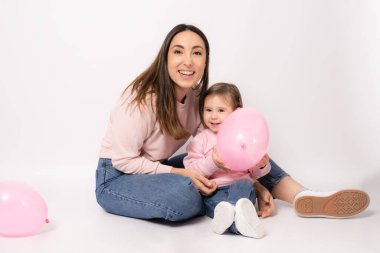 Küçük kızı olan mutlu bir anne beyaz arka planda pembe balonlarla yerde oturuyor..