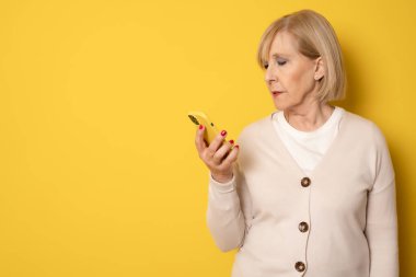 Gündelik kıyafet konuşmalarında 70 'li yaşlarda gülümseyen mutlu bir kadın sarı arka plan stüdyo resminde izole edilmiş cep telefonuyla konuşuyor. Halk yaşam tarzı konsepti.