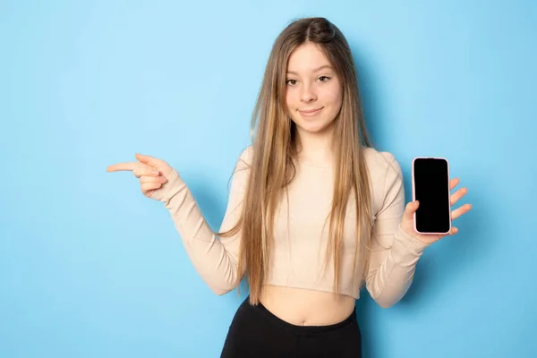 スタイリッシュな女の子は空の携帯電話の画面を表示し 喜んで見て スマートフォンのためのアプリケーションをお勧めします 青の背景にショッピングサイトを実証 — ストック写真