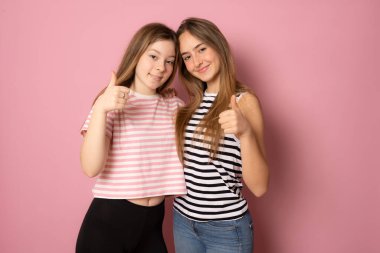 İki neşeli genç kadının portresi yan yana duruyor ve pembe arka planda parmakları yukarıda kameraya bakıyor.