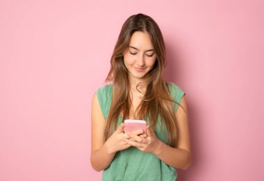 Pembe arka planda izole edilmiş cep telefonuna bakan gülümseyen, sıradan, beyaz bir kadının portresi.