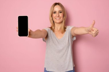 Mutlu, güzel, günlük kıyafetler giyen, pembe arka planda boş ekranlı cep telefonu tutan bir kadın.