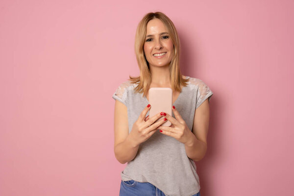 Портрет улыбающейся молодой кавказки, смотрящей на мобильный телефон на розовом фоне
