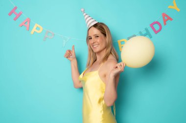 Sarı balon ve pastayla doğum gününü kutlayan gülümseyen kadın, şenlik şapkası takıyor, arkadaşlarının sürprizinin tadını çıkarıyor, yeşil arka planda izole edilmiş.