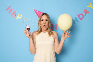 Sarı balon ve pastayla doğum gününü kutlayan gülümseyen kadın, şenlikli şapka takıyor, arkadaşlarının sürprizinin keyfini çıkarıyor, mavi arka planda izole edilmiş.