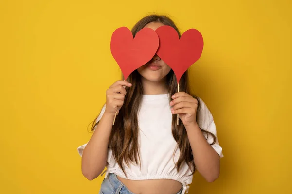 Girl covering eyes with two carton paper small little heart figures standing isolated over yellow background. Love concept.